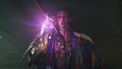 A person in dramatic, dark lighting holds a glowing purple sword or staff while wearing layered metallic necklaces and a colorful patterned garment. The scene is moody and cinematic, with a blurred dark background.