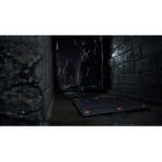 A dark, narrow stone corridor with a black rectangular floor mat or panel laid on the ground, shown in a dimly lit environment.